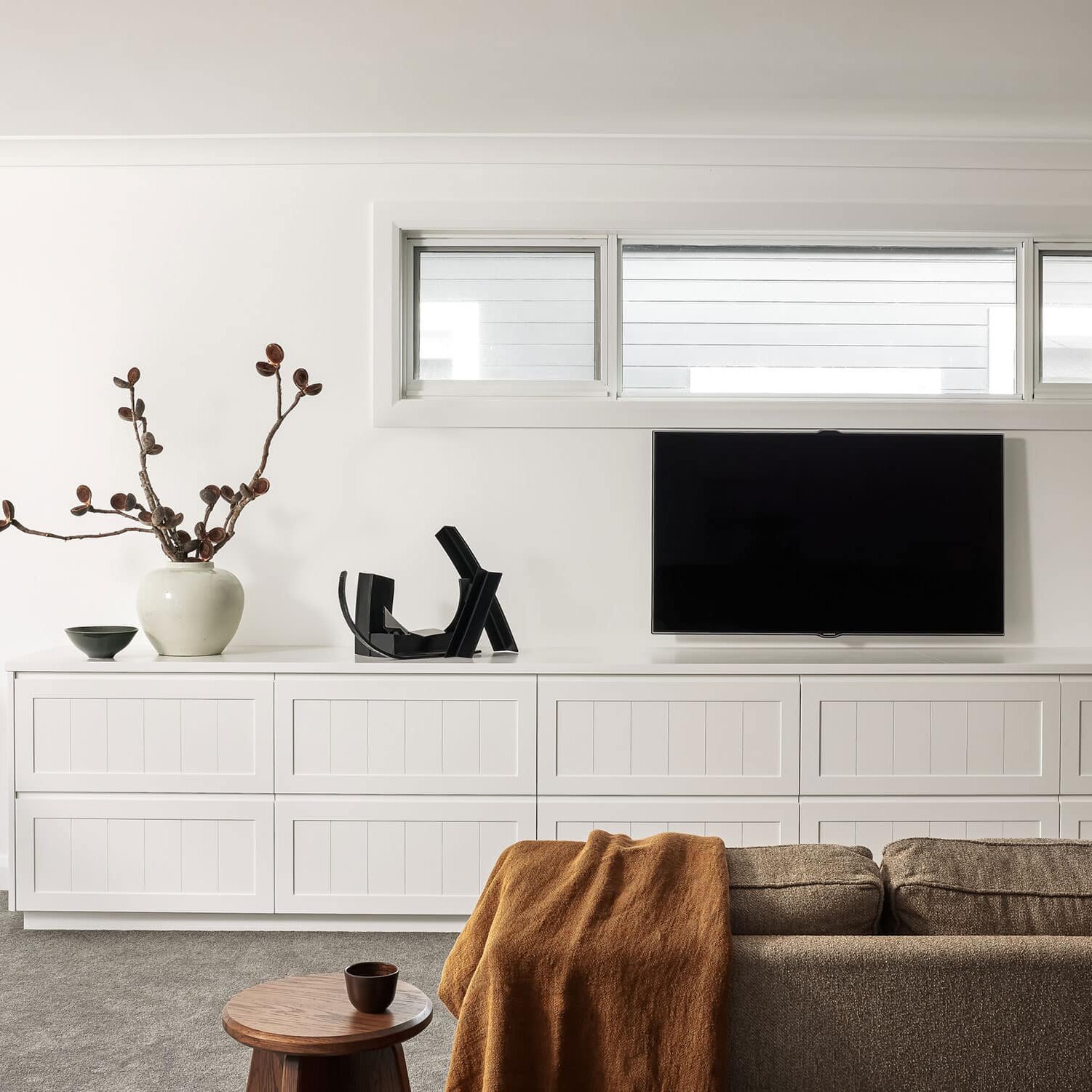 Bright living room with white built-in cabinets, a wall-mounted TV, and a brown sofa with a rust-colored throw.