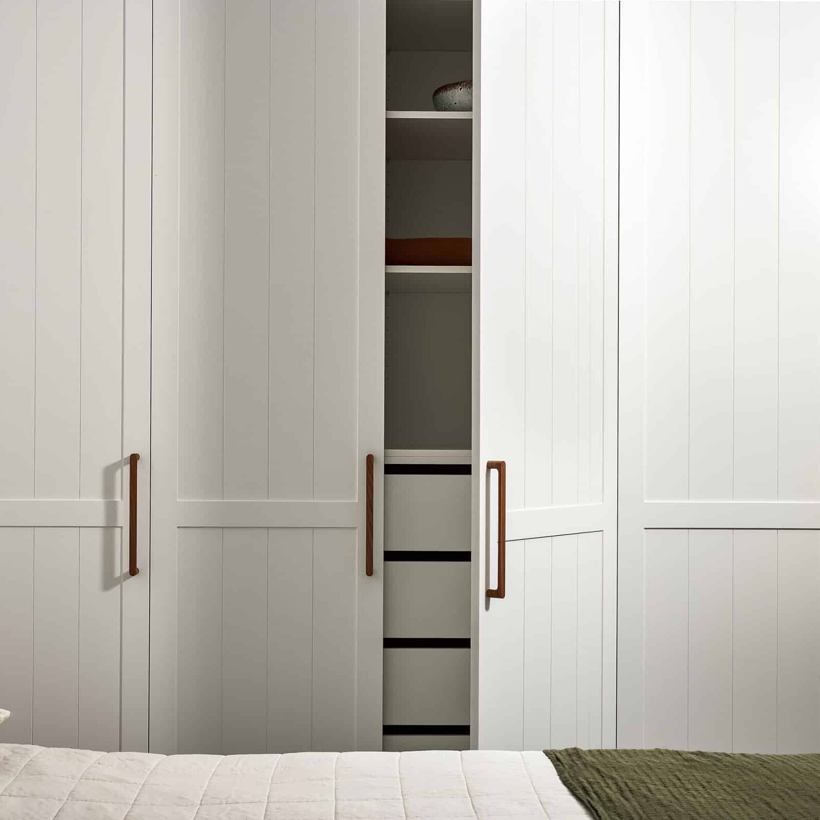 White built-in wardrobe doors with vertical panels and wooden handles; the center section is open, revealing shelves with a ceramic bowl on the top shelf and a folded brown towel on a lower shelf