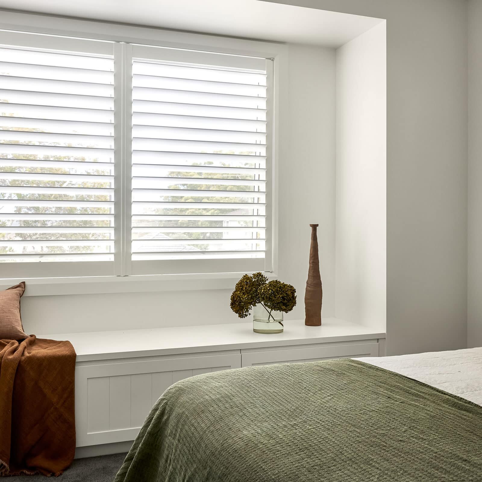 Bedroom with a large window and white horizontal blinds above a white window seat; olive-green bedspread in foreground and an orange throw draped over the seat.